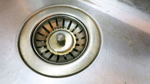 Abflusssieb mit Backofenspray einsprühen und ein paar Minuten einwirken lassen Ein Abflusssieb in einer Küchenspüle, das zum Reinigen mit Backofenspray vorbereitet ist, zeigt Ablagerungen.