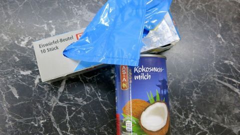Eine Dose Kokosmilch steht auf einem dunklen Marmortisch. Ein blauer Eiswürfel-Beutel mit Verpackung liegt daneben. Text: "Eiswürfel-Beutel 10 Stück".