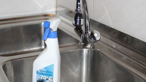 Fingerabdrücke oder andere Verschmutzungen auf Edelstahlteilen lassen sich mit Scheibenenteiser entfernen. Ein Spray zur Scheibenenteiser für Edelstahlglanz steht neben einem glänzenden Wasserhahn in einer modernen Küche.
