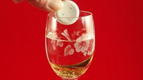 Ein Finger taucht eine Reinigungstablette in ein elegantes Glas mit Wasser und goldfarbenen Bläschen vor rotem Hintergrund ein.