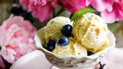 Eine dekorative Schale mit Eiscreme wird von blühenden Pfingstrosen umgeben, während einige Heidelbeeren darauf liegen.