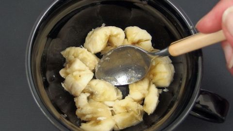 Eine Hand benutzt einen Löffel, um zerdrückte Bananen in einer schwarzen Tasse zu vermischen. Kein Ei im Haus zum Kuchen backen?