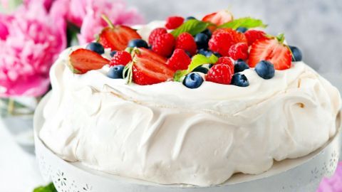 Eine Pavlova mit frischen Früchten steht auf einem eleganten Tisch. Bunte Beeren und Erdbeeren dekorieren die luftige Süßigkeit.
