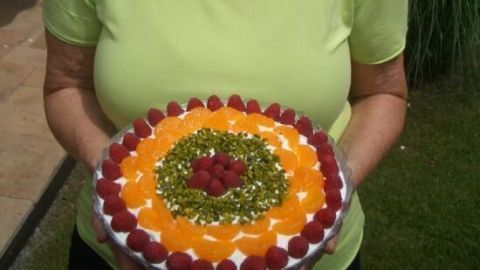 Eine Frau hält stolz einen bunten Sherry Himbeer Trifle in einem Garten, umgeben von grünen Pflanzen und Sonne.