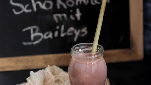 Ein Glas warme Schokomilch mit Baileys steht auf einem Tisch, umgeben von einem Tuch, vor einer beschrifteten Tafel.