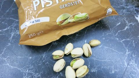 Gehackte Pistazien liegen auf einer dunklen Oberfläche neben einer offenen Tüte. Die Pistazien sind in der Tüte sichtbar.