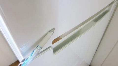 Mit Nagellackentferner Kugelschreiberflecken von Kühlschranktüren entfernen. Ein Kugelschreiber liegt auf einer Kühlschranktür, während er einen Fleck entfernt. Der Raum ist hell und minimalistisch.
