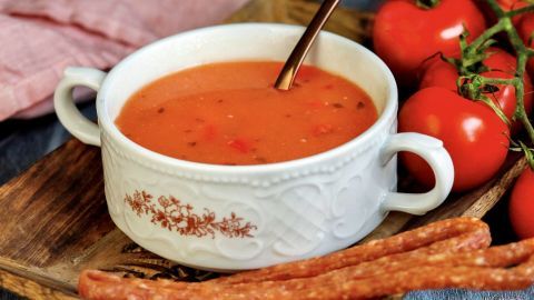 Eine bunte Schüssel mit Tomatensuppe steht auf einem Holzbrett, umgeben von frischen Tomaten und knusprigen Stangen.