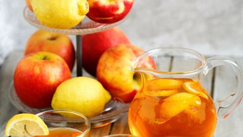 Zwei Gläser mit orangefarbenem Getränk stehen vor einem Krug. Dahinter liegen frische Äpfel und Zitronen auf einem Ständer.