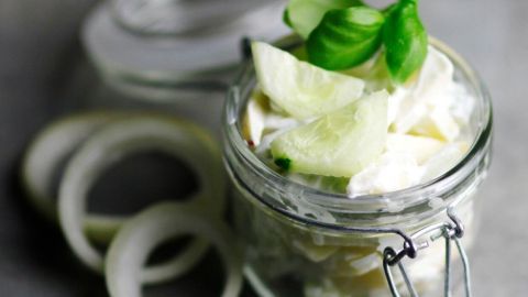 Eine Glasbehälter mit Gurkensalat und Apfelstücken wird durch frische Basilikumblätter dekoriert, umgeben von Zwiebelringen.