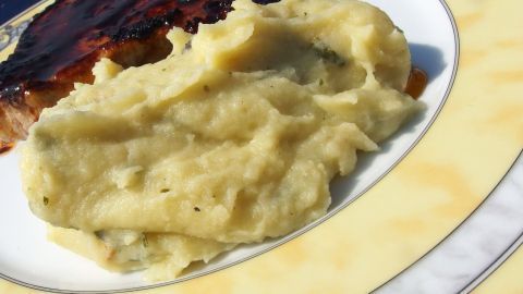 Eine Schüssel Kartoffelbrei liegt auf einem Teller neben einem Stück Fleisch, appetitlich angerichtet im Freien.