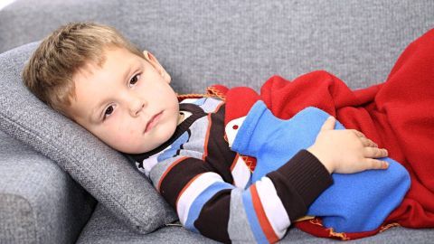Ein Kind liegt auf einem Sofa, hält eine blaue Wärmflasche auf dem Bauch und ist mit einer roten Decke zugedeckt.