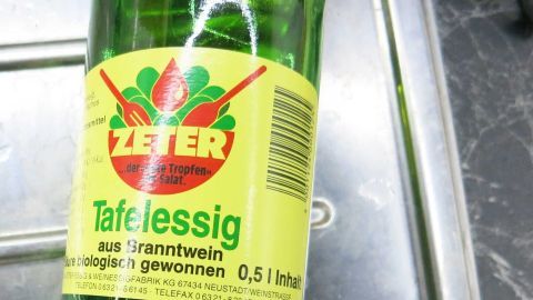 Tafelessig ist ein effektives Mittel, um Grünspan im Spülbecken zu entfernen. Gieße etwas Essig auf die betroffene Stelle, lass ihn einwirken und wische dann mit einem Tuch nach. So wird dein Spülbecken wieder strahlend sauber. Essigflasche steht im Spülbecken. Das Etikett zeigt: "ZETER Tafelessig, aus Branntwein, 0,5 l, biologisch gewonnen".