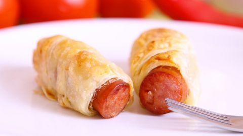 Zwei Würstchen in Blätterteig liegen auf einem weißen Teller, umgeben von roten Tomaten und Pfeffer.