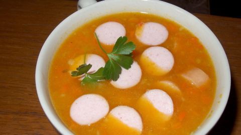 Eine Portion Kürbissuppe, angereichert mit Würstchen, ist in einer weißen Schüssel serviert und mit frischer Petersilie garniert.