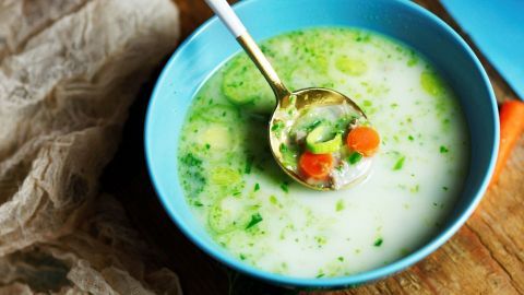 Eine goldene Löffel hebt eine cremige Möhren-Porree-Hackfleisch-Suppe in einer blauen Schüssel, umgeben von frischem Gemüse.