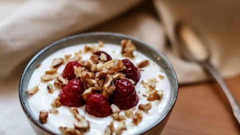 Eine Schüssel Joghurt mit frischen Himbeeren und gehackten Nüssen steht auf einem Tisch, umgeben von einem weichen Tuch.