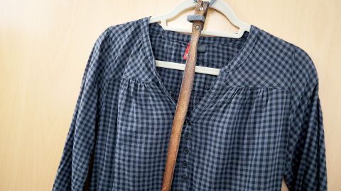 Eine lange Bluse hängt an einem Kleiderbügel neben einem Stock an einer hellen Wand, bereit zum Tragen oder Stylen.