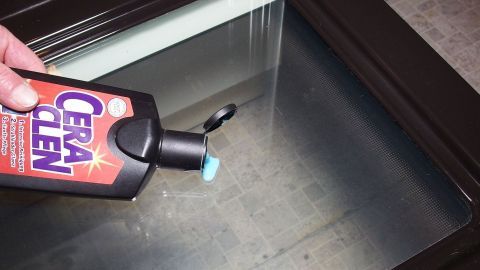 Eine Hand hält eine Flasche Glaskeramikreiniger über ein Backofensichtfenster und bereitet das Reinigen vor.