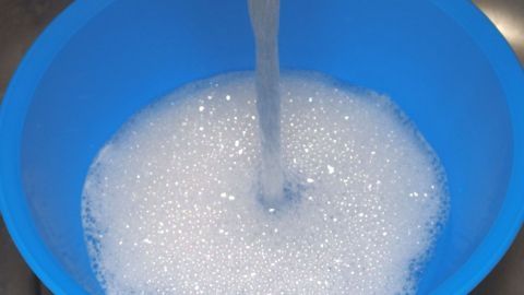 Beim Spülen von Hand Wasser sparen - spülen in einer Plastikschüssel. Ein blauer Eimer wird mit Wasser gefüllt, während sich Schaum oben bildet, um beim Spülen von Hand Wasser zu sparen.