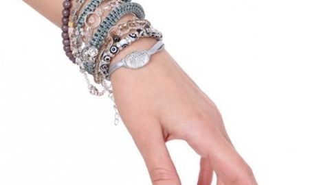 Eine Hand mit mehreren Armbändern und Armkettchen, locker hängend, vor einem weißen minimalistischen Hintergrund.