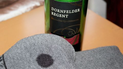 Ein graues Textilstück mit einem Rotweinfleck liegt neben einer Flasche Dornfelder, die auf einem Tisch steht.