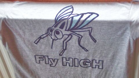 Ein T-Shirt mit einem großen, cartoonhaften Fliegenmotiv und dem Text „Fly HIGH“ hängt an einer Wäscheleine.