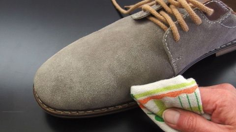 Schneeränder auf Wildlederschuhen entfernen mit destilliertem Wasser. Ein grauer Lederschuh wird mit einem feuchten Tuch gereinigt, um Schneeränder zu entfernen, auf einem dunklen Tisch.