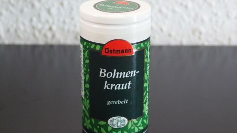 Ein Behälter mit Bohnenkraut steht auf einem dunklen Tisch, während der Hintergrund eine helle Wand zeigt.