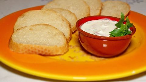 Auf einem gelben Teller liegt frisches Brot, daneben ein rotes Schälchen mit einem cremigen Frischkäse-Schinken-Aufstrich.