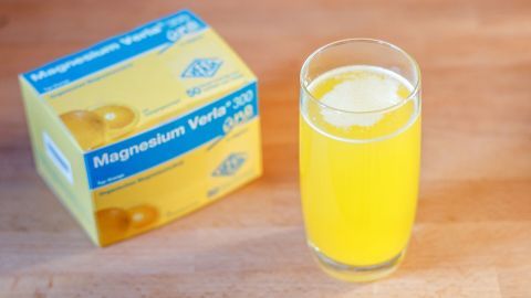 Ein Glas mit gelbem Getränk, vermutlich Magnesium, steht neben einer Schachtel "Magnesium Verla 300" auf einem Holztisch.