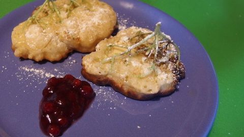 Zwei gebackene Holunderblüten auf einem blauen Teller, mit Puderzucker bestreut, neben einer Portion Himbeermarmelade.