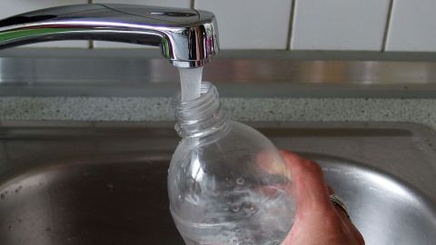 Eine PET-Flasche wird unter einem Wasserhahn gefüllt, während die Hand sie hält, in einer Küche mit Fliesen.