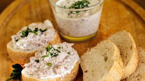 Auf einem breiten Holzbrett liegen Scheiben Brot mit cremiger Thunfischcreme und ein Glas mit Dip, umgeben von frischen Kräutern.
