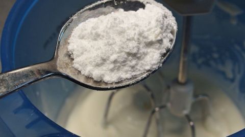 Sahne bleibt bleibt länger steif wenn man sie mit Puderzucker süßt. Ein Löffel mit Zucker schwebt über einer Schüssel mit geschlagener Sahne, die mit einem Mixer zubereitet wird.