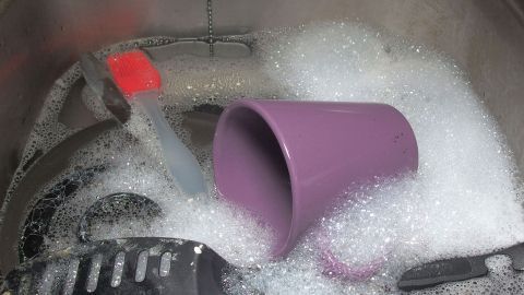 Ein lila Becher liegt schräg in einer Spülmaschine, umgeben von Schaum und Geschirr, während die Klappe der Maschine offen ist.
