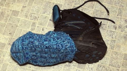Eine schwarze Glatteisbremse liegt auf einem hellen, strukturierten Untergrund, während daneben ein blauer, gestrickter Teil sichtbar ist.