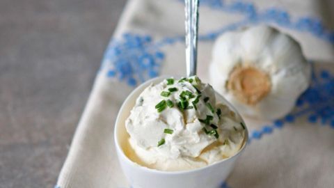 Ein cremiger Knoblauchdip in einer weißen Schale mit frischen Kräutern, neben einer Knoblauchzehe auf einem Tisch.
