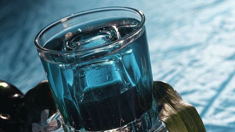 Ein Glas mit blauem Getränk steht auf einem gläsernen Untersetzer, umgeben von grünen Blättern auf einem blauen Tisch.