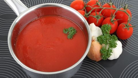 Eine silberne Kasserolle mit roter Tomatensauce steht auf einer Arbeitsfläche, umgeben von frischen Tomaten, Zwiebeln und Knoblauch.