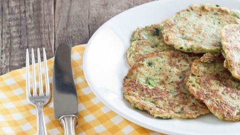 Zucchinipuffer auf weißem Teller liegen auf rustikalem Holztisch neben einem gelben Tischset, Zucchini im Hintergrund.