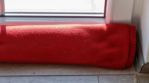 Ein roter Zugluftstopper liegt am Fenster, um unerwünschte Zugluft im Raum zu verhindern, während Sonnenlicht einfällt.