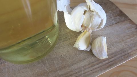 Eine Flasche mit aromatisiertem Öl und frischen Knoblauchzehen liegen auf einem Holzbrett in einer hellen Umgebung.