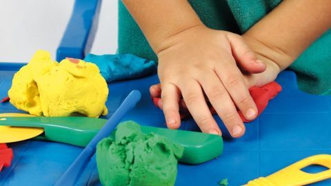 Ein Kind formt Knetmasse mit seinen Händen auf einem blauen Tisch, umgeben von bunten Knetfarben und Werkzeugen.