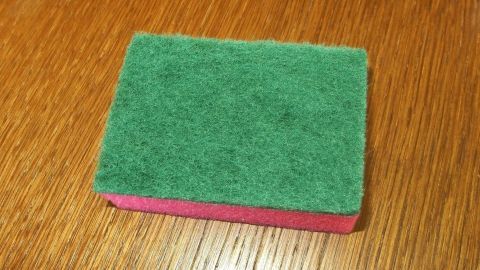 Zur Entfernung von Hornhaut an den Füßen eignet sich ein Cellulose-Schwamm mit Scheuervlies sehr gut. Ein Scheuerschwamm liegt auf einem Holzuntergrund. Er wird zum Entfernen von Hornhaut an Füßen verwendet.