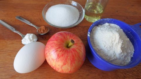 Ein Apfel, ein Ei und Mehl in einer blauen Schale liegen auf einem Holztisch, begleitet von Zucker, Öl, Zimt und Backpulver.
