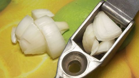 Fein zerkleinerte Zwiebeln liegen neben einem Zwiebelhacker auf einer bunten Unterlage mit frischen Früchten.
