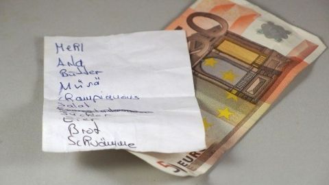 Eine Einkaufsliste liegt neben einem 10-Euro-Schein auf einem Tisch. Die Liste enthält verschiedene Lebensmittel und Artikel.