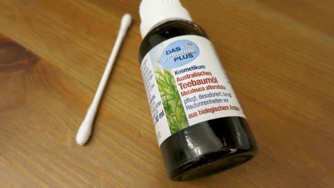 Eine Flasche "Australisches Teebaumöl" liegt neben einem Wattestäbchen auf einem Holztisch. Text auf der Flasche: "Kosmetikum Australisches Teebaumöl Melaleuca alternifolia pflegt, desodoriert, beugt Hautunreinheiten vor aus biologischem Anbau".