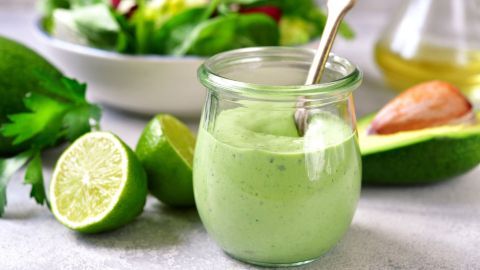 Ein hellgrünes Joghurt-Pesto-Dressing steht in einem Glas, während Limettenhälften und Avocado daneben liegen, umgeben von frischem Salat.